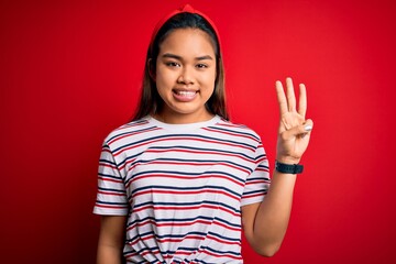 Young beautiful asian girl wearing casual striped t-shirt over isolated red background showing and pointing up with fingers number three while smiling confident and happy.