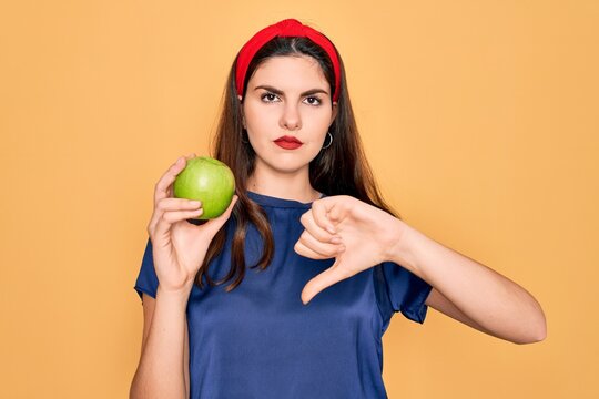 Young beautiful girl eating fresh organic healthy green apple over yellow background with angry face, negative sign showing dislike with thumbs down, rejection concept
