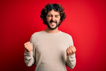 Young handsome man with beard wearing casual sweater standing over red background very happy and excited doing winner gesture with arms raised, smiling and screaming for success. Celebration concept.