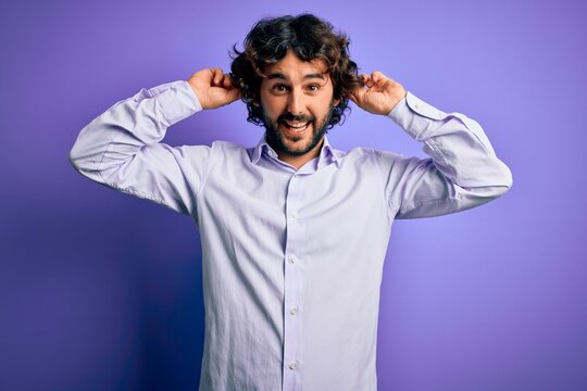 Young handsome business man with beard wearing shirt standing over purple background Smiling pulling ears with fingers, funny gesture. Audition problem