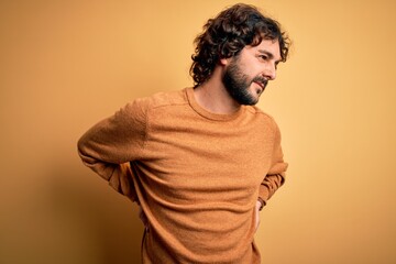 Young handsome man with beard wearing casual sweater standing over yellow background Suffering of backache, touching back with hand, muscular pain