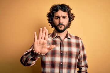 Young handsome man with beard wearing casual shirt standing over yellow background doing stop sing...