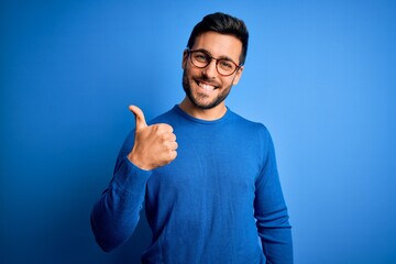 Young handsome man with beard wearing casual sweater and glasses over blue background doing happy thumbs up gesture with hand. Approving expression looking at the camera showing success. - obrazy, fototapety, plakaty