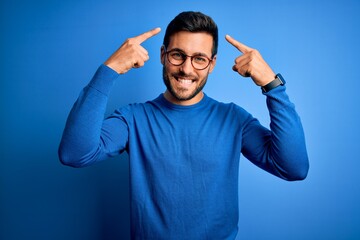Young handsome man with beard wearing casual sweater and glasses over blue background smiling...