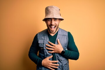 Handsome tourist man with beard on vacation wearing explorer hat over yellow background smiling and laughing hard out loud because funny crazy joke with hands on body.