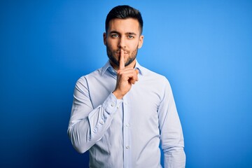 Young handsome man wearing elegant shirt standing over isolated blue background asking to be quiet with finger on lips. Silence and secret concept.