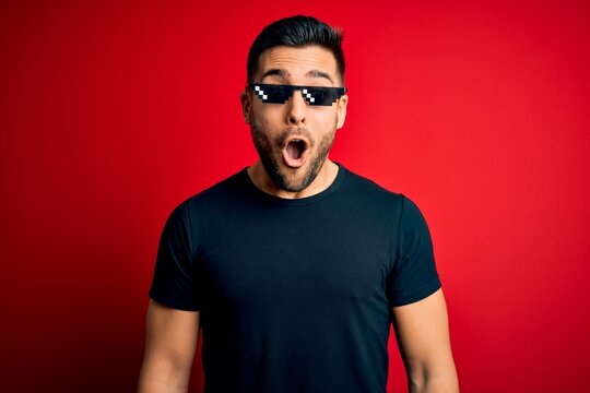 Young handsome man wearing funny thug life sunglasses over isolated red background afraid and shocked with surprise expression, fear and excited face.