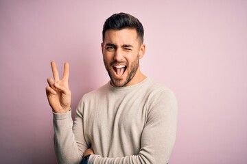 Young handsome man wearing casual sweater standing over isolated pink background smiling with happy face winking at the camera doing victory sign. Number two.