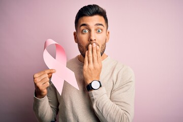 Young handsome man holding pink cancer ribbon symbol of support over isolated background cover mouth with hand shocked with shame for mistake, expression of fear, scared in silence, secret concept