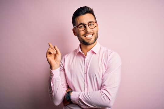 Young handsome man wearing elegant shirt and glasses standing over pink background with a big smile on face, pointing with hand and finger to the side looking at the camera.