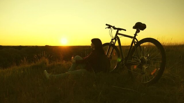 Tired woman cyclist resting in evening on edge of a cliff and admiring sun. Free girl travels with bicycle at sunset. Healthy young woman hiker sitting on hill next to bicycle, enjoying nature and sun