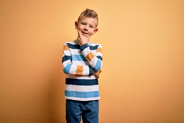 Young little caucasian kid with blue eyes wearing colorful striped shirt over yellow background looking confident at the camera smiling with crossed arms and hand raised on chin. Thinking positive.