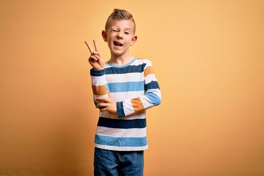 Young little caucasian kid with blue eyes wearing colorful striped shirt over yellow background smiling with happy face winking at the camera doing victory sign. Number two.