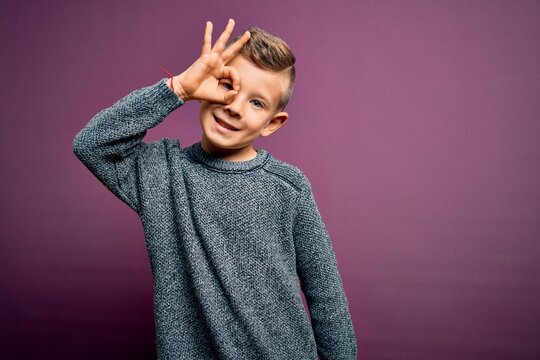 Young little caucasian kid with blue eyes wearing winter sweater standing over purple background doing ok gesture with hand smiling, eye looking through fingers with happy face.