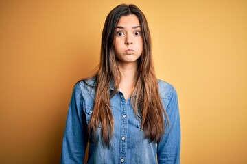 Young beautiful girl wearing casual denim shirt standing over isolated yellow background puffing cheeks with funny face. Mouth inflated with air, crazy expression.