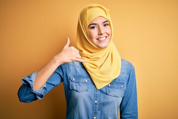 Young beautiful girl wearing muslim hijab standing over isolated yellow background smiling doing phone gesture with hand and fingers like talking on the telephone. Communicating concepts.