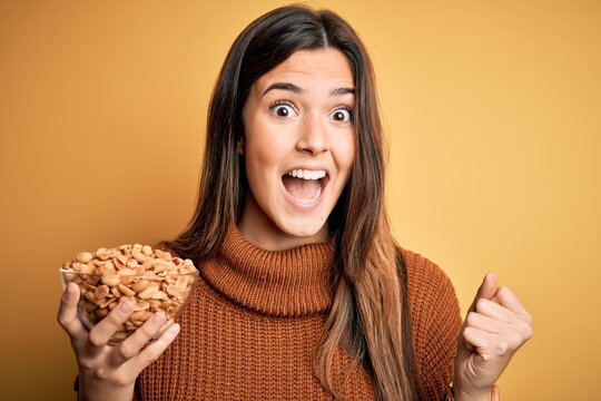 Young beautiful girl holding bowl of salty peanuts standing over yellow background screaming proud and celebrating victory and success very excited, cheering emotion