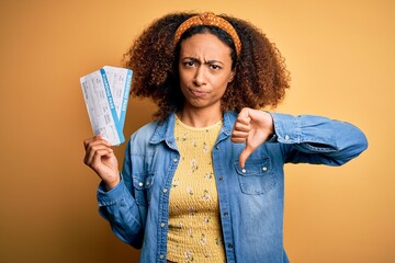 Young african american woman with afro hair holding plane boarding pass for a holiday trip with angry face, negative sign showing dislike with thumbs down, rejection concept