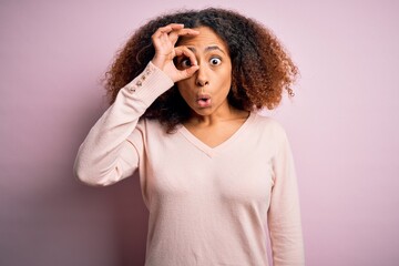 Young african american woman with afro hair wearing casual sweater over pink background doing ok gesture shocked with surprised face, eye looking through fingers. Unbelieving expression.