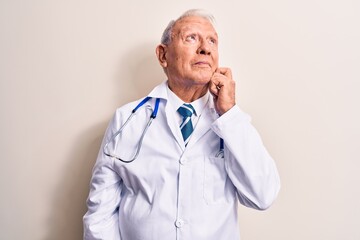 Senior grey-haired doctor man wearing coat and stethoscope standing over white background thinking concentrated about doubt with finger on chin and looking up wondering