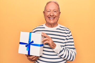 Senior handsome grey-haired man holding birthday gift over isolated yellow background looking positive and happy standing and smiling with a confident smile showing teeth