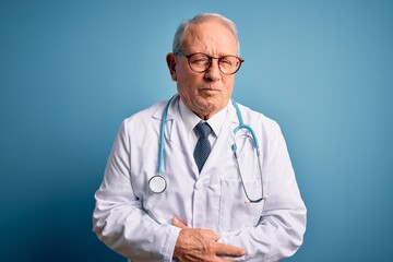 Senior grey haired doctor man wearing stethoscope and medical coat over blue background with hand on stomach because indigestion, painful illness feeling unwell. Ache concept.