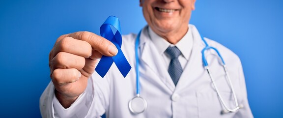 Grey haired senior doctor man holding colon cancer awareness blue ribbon over blue background with a happy face standing and smiling with a confident smile showing teeth