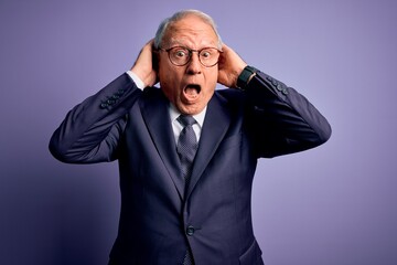 Grey haired senior business man wearing glasses and elegant suit and tie over purple background Crazy and scared with hands on head, afraid and surprised of shock with open mouth