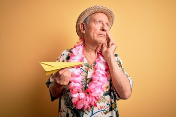 Grey haired senior man wearing summer hat and hawaiian lei holding paper plane on vacation serious face thinking about question, very confused idea
