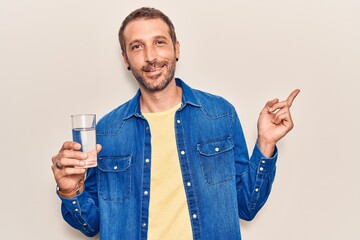 Young handsome man drinking glass of water smiling happy pointing with hand and finger to the side