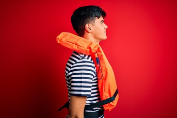 Young handsome man wearing orange safety life jacket over isolated red background looking to side, relax profile pose with natural face with confident smile.