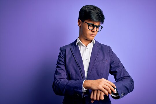 Young handsome business man wearing jacket and glasses over isolated purple background Checking the time on wrist watch, relaxed and confident