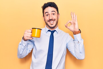 Young hispanic man wearing business clothes holding coffee doing ok sign with fingers, smiling friendly gesturing excellent symbol