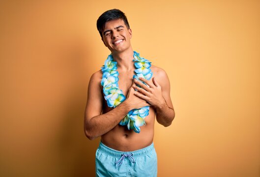 Young handsome tourist man on vacation wearing swimwear and hawaiian lei flowers smiling with hands on chest with closed eyes and grateful gesture on face. Health concept.