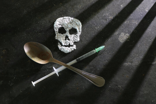 Heroin cooking spoon and hypodermic syringe below a white powder skull. Skull and cross bones image. Shadows imply prison bars. Drug use in prisons concept. Intentionally underexposed for mood.