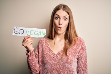 Young beautiful redhead woman asking for veggie food holding paper with vegan message scared in...