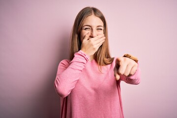 Young beautiful redhead woman wearing casual sweater over isolated pink background laughing at you, pointing finger to the camera with hand over mouth, shame expression