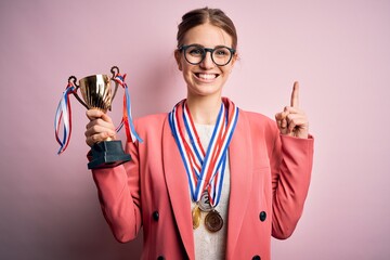 Young beautiful redhead woman holding trophy wearing medals over pink background surprised with an...