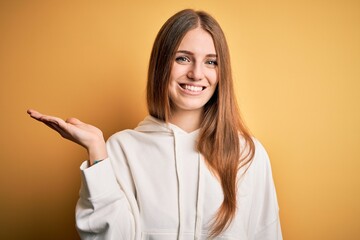 Young beautiful redhead sporty woman wearing sweatshirt over isolated yellow background smiling cheerful presenting and pointing with palm of hand looking at the camera.