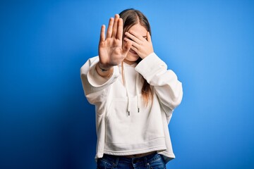 Young beautiful redhead sporty woman wearing sweatshirt over isolated blue background covering eyes with hands and doing stop gesture with sad and fear expression. Embarrassed and negative concept.