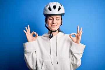 Young beautiful redhead cyclist woman wearing bike helmet over isolated blue background relax and smiling with eyes closed doing meditation gesture with fingers. Yoga concept.