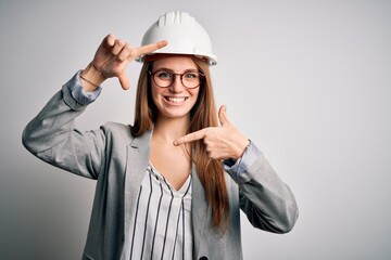 Young beautiful redhead architect woman wearing security helmet over white background smiling making frame with hands and fingers with happy face. Creativity and photography concept.