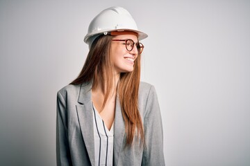 Young beautiful redhead architect woman wearing security helmet over white background looking away to side with smile on face, natural expression. Laughing confident.
