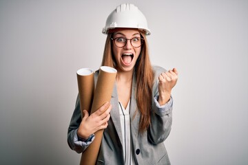 Young beautiful redhead architect woman wearing glasses and safety helmet holding blueprints screaming proud and celebrating victory and success very excited, cheering emotion