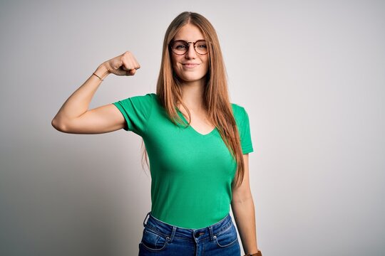 Young beautiful redhead woman wearing casual green t-shirt and glasses over white background Strong person showing arm muscle, confident and proud of power