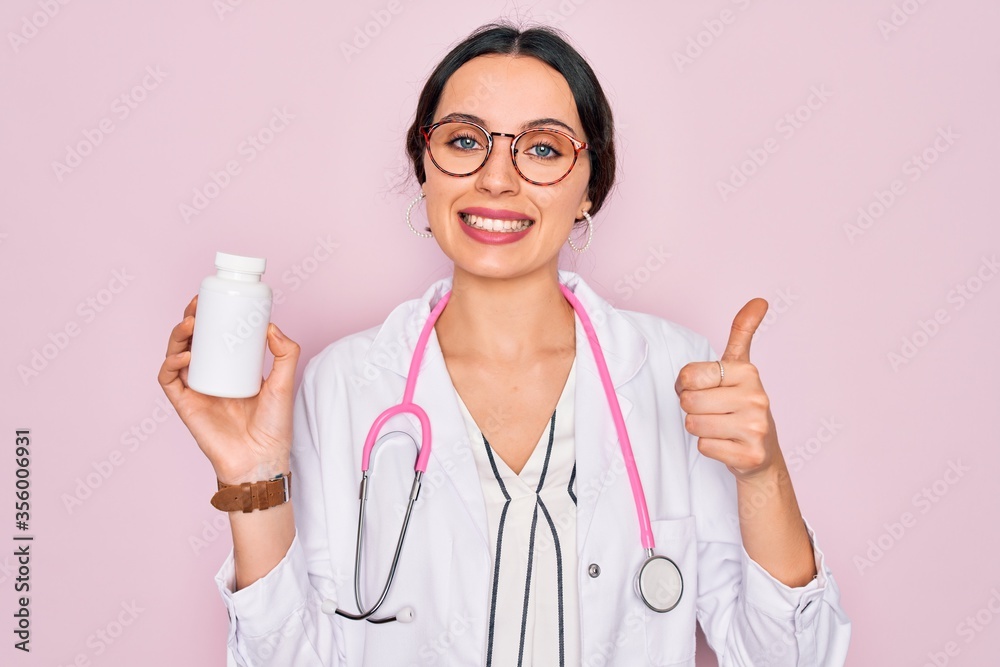 Wall mural Young beautiful doctor woman with blue eyes wearing stethoscope holding medicine pills happy with big smile doing ok sign, thumb up with fingers, excellent sign