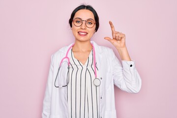 Beautiful doctor woman with blue eyes wearing coat and stethoscope over pink background smiling and confident gesturing with hand doing small size sign with fingers looking and the camera. Measure