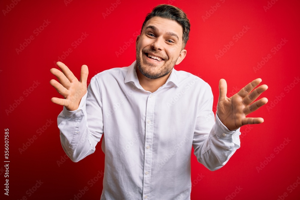 Wall mural young business man with blue eyes wearing elegant shirt standing over red isolated background celebr