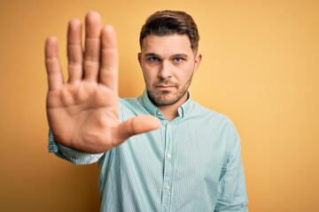 Young business man with blue eyes wearing elegant green shirt over yellow background doing stop sing with palm of the hand. Warning expression with negative and serious gesture on the face.