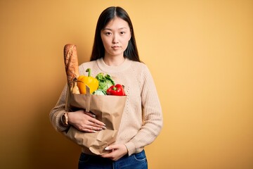 Young asian woman holding paper bag of fresh healthy groceries over yellow isolated background Relaxed with serious expression on face. Simple and natural looking at the camera.